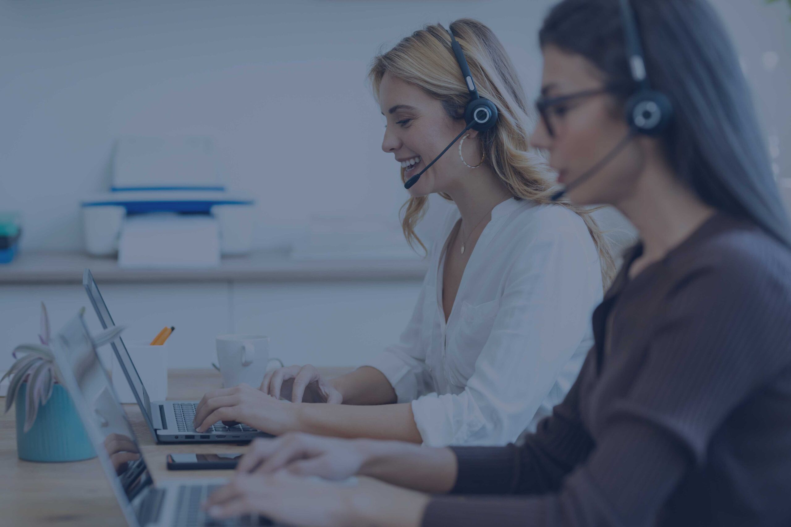 Due addette al customer service, sedute davanti al computer, rispondo alle chiamate tramite le loro cuffie.