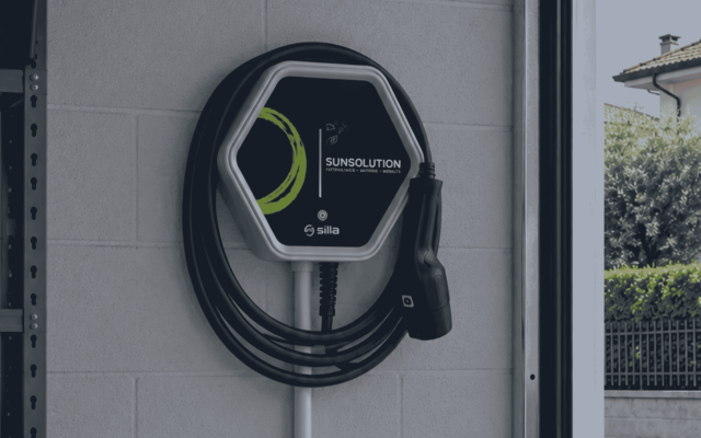 Vista dal basso di un'auto nera parcheggiata sotto a un carport fotovoltaico, attaccata a una wallbox per il caricamento della batteria.