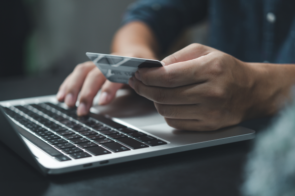 Una persona a mezzo busto è seduta al tavolo con davanti il proprio pc portatile: con una mano tiene la carta di credito, con l'altra sta digitando la tastiera del pc.
